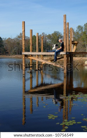 Man sits on wooden dock.  His dog is keeping him company.  Reflection of both are in the still waters of the lake.  Blue skies and blue water with lilly pads.