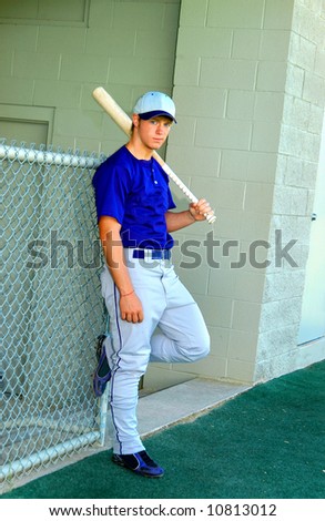 Young male teen leans against dugout wall as he waits to his turn at bat.  He is holding a bat and one foot leans against metal fence.