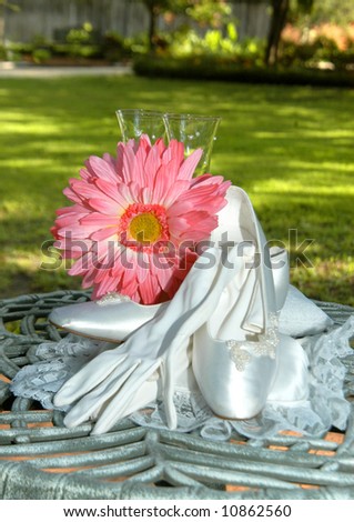 Wedding accessories are arranged on garden table.  Hot pink daisy sits in front of wine goblet.  Satin shoes and gloves.