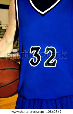 Blue basketball jersey is worn by male teen in basketball game.  Basketball is under his arm.
