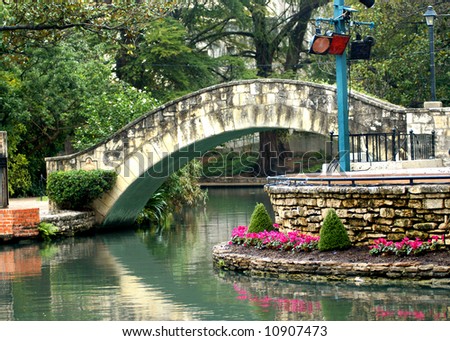 Bridge is reflected in the still waters of river on the San Antonio River Walk.  Flowers in pink bloom along river\'s edge.