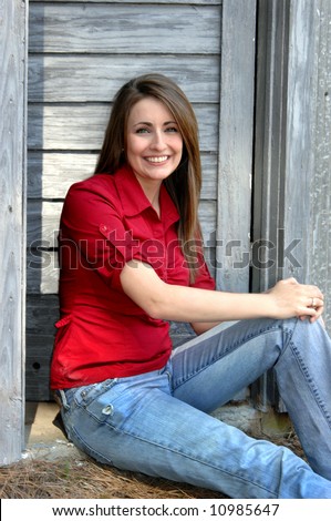 Young woman sits at the door of an old wooden barn.  She is wearing a red shirt and jeans.  She is smiling.