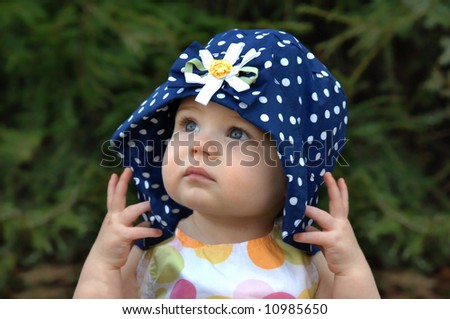 Baby girl poses with a floppy hat.  Hat is navy with white polka dots.  Solemn expression.