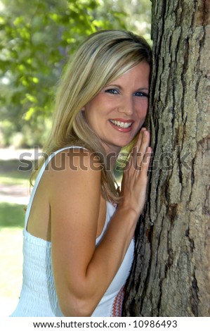 Beautiful young woman leans against a tree at a park.  Sleeveless dress.  She is smiling happily.