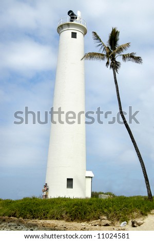 Barber\'s Point lighthouse has tropical setting.  One lone palm tree.  Couple lean against base of lighthouse.