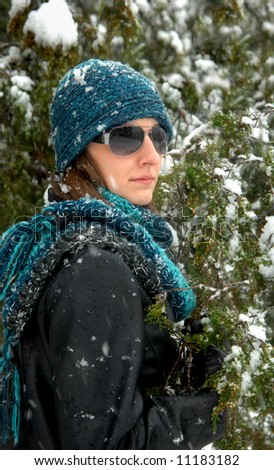 Winter snowflakes fall on young woman and fur trees.  Black leather jacket, blue and turquoise knit cap and scarf.  Sunshades.