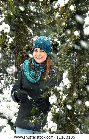 Winter snowflakes fall on young woman and fur trees.  Black leather jacket, blue and turquoise knit cap and scarf.  Smile.