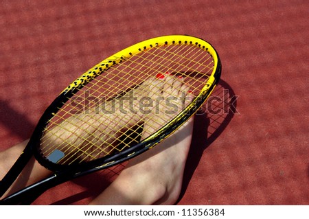 Tennis toes peek out from beneath a tennis racket.  Bare feet with red toe nails.  Red paved court.