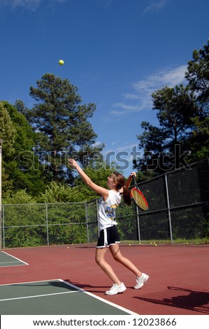 Energetic teen serves tennis ball during a high school tennis match.  Uniform is black skirt and white tee shirt.