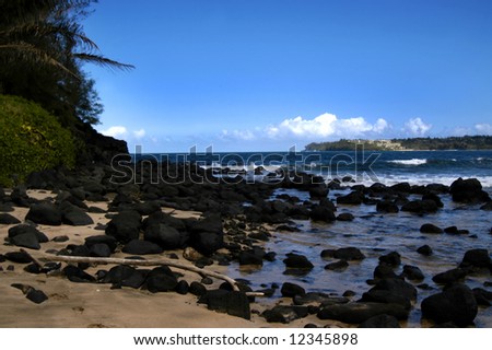 Black rocks fill beach in Hanalei Bay on Kauai, Hawaii.  Deep aqua blue water with vivid blue sky.