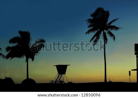 Beginning of a new day for lifeguard.  Shack and palm trees are silhouetted as the first light of day colors the sky orange and yellow.