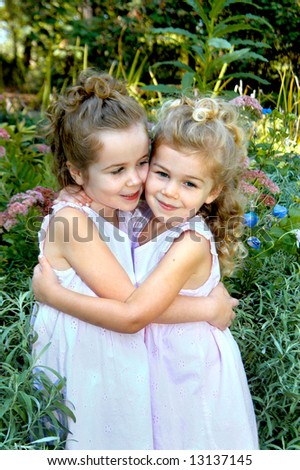 Best friends hug each other and smile.  Outdoors in park with flowers and sunshine in background.  One sister is looking at the other one.