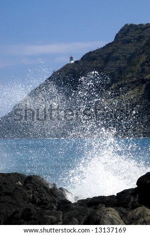 Distant Makapu\'u Head Lighthouse is framed by wildly splashing wave.  Blue skies and water.