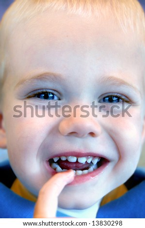 A big smile from a sweet small boy with big blue eyes and blonde hair.  He has one finger in his mouth.  Blue shirt.