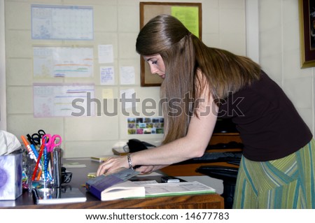 Female office worker makes a list of things-to-do that day.  She has long hair, patterned skirt and brown shirt.  Working at desk.