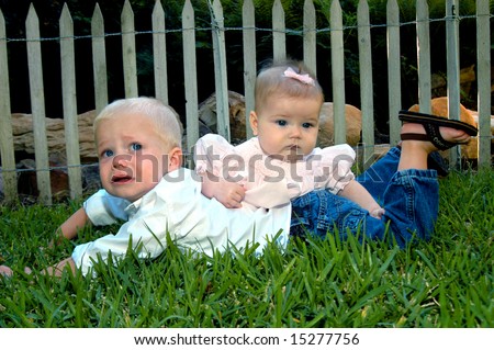 Brother is not happy about his sister\'s affection.  Baby girl lays across small boys back.  Both are laying on the grass.