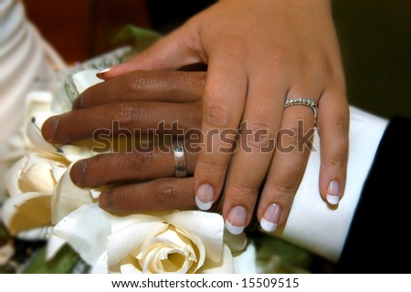 Man and woman of mixed race unite in marriage.  Hands with wedding bands lay over bouquet.