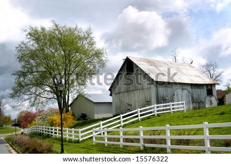 Springtime surrounded country barn.  White picket fence surrounds barn and pasture.  City street runs besides fence.