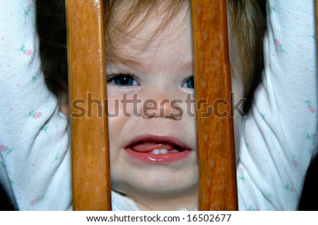 Small female toddler peeks through the slats of her crib.  She is afraid to go to bed.