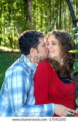 Young couple embrace in garden.  He is kissing her on the cheek.