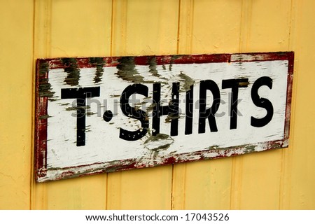 Rustic T-Shirts sign is weathered and its paint is peeling.  It is mounted on a yellow-gold wooden building.