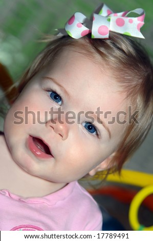 Baby girl stares with open mouth.  She has on pink including hair bow in colorful dots.  Big blue eyes.