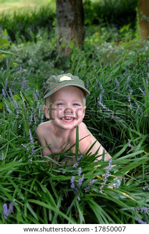 Small boy grins from ear to ear as he poses in a garden.  Green army cap.