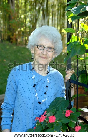 Elegant grandmother poses besides a black iron fence covered in ivy and flowers.  Blue jacket and matching sweater.