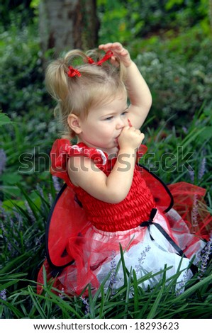 Baby girl dressed as a lady bug in costume with wings and net, sits in garden and sucks her thumb.