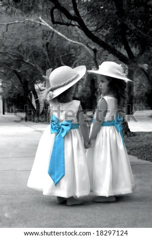 Beautiful young girls in white dresses with blue bows walk holding hands in a garden.  They are wearing hats and photo is black and white.