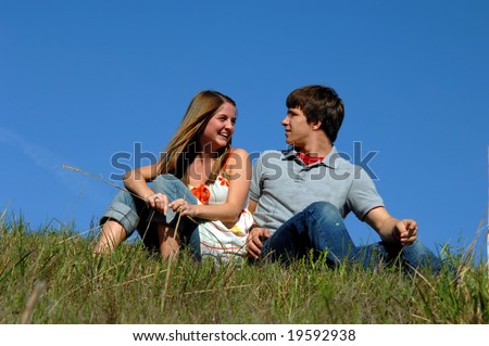 Blue skies frame young teen couple.  They are sitting on a grassy hill side-by side.