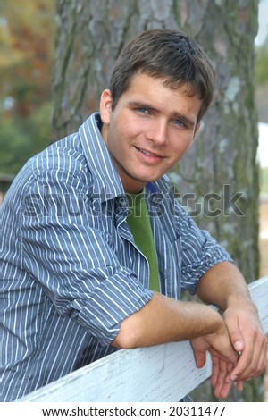 Attractive male teen leans over white wooden fence.  He is wearing long sleeved dress shirt and smiling.