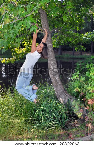 Kicking her heels up, a female teen swings on the limb of a tree besides a small pond.