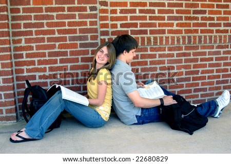 Back to back, two students study while sitting on a sidewalk.  The teenage girl is smiling and the boy is reading.
