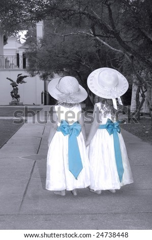 Two small girls walk down sidewalk in their long dresses with blue bows and wide brim hats.  Black and white colorized with blue bows.