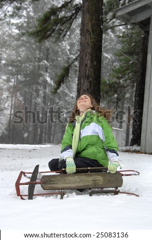 Falling snow and sledding makes this young woman laugh in enjoyment as she sits in the snow with wooden sled.