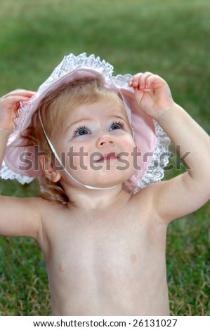 Sweet little girl models a frilly and lacy pink hat with elastic band under her chin.  She is looking up and is unsmiling.