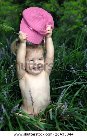 Female child sits in a bed of tall green grass and plays peek a boo with her hot pink cap.
