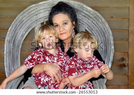 Grandmother hugs her twin grandsons up close.  Boys are wearing identical shirts and shorts.  Grandmother is wearing black and jeans.