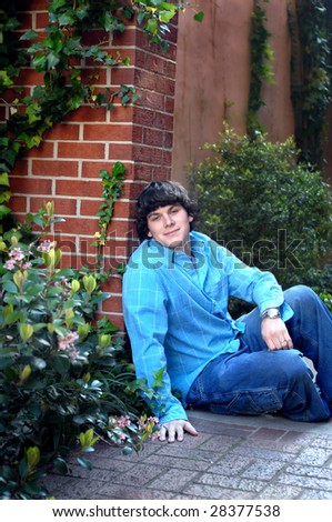 Young man leans against a brick wall in a garden.  He is wearing jeans and a blue shirt.