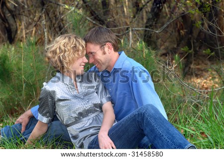 Affectionate couple touch foreheads as they sit in grass outdoors.  Woods and trees fill background.