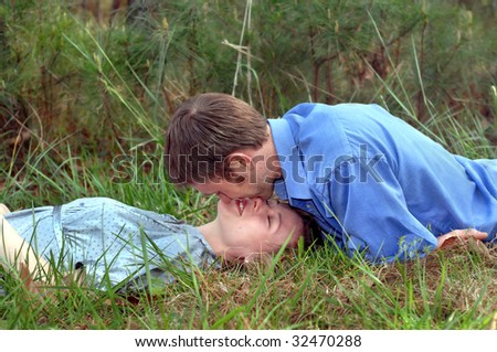 Couple share a romantic kiss laying on the grass in woods.  He is wearing a blue shirt and she a grey silk blouse.