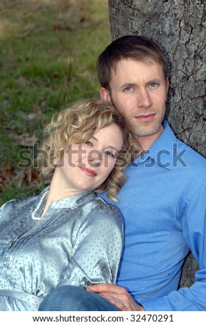 Couple sit close together against a tree trunk outdoors.  He is wearing a blue shirt and jeans and she a grey silk blouse.