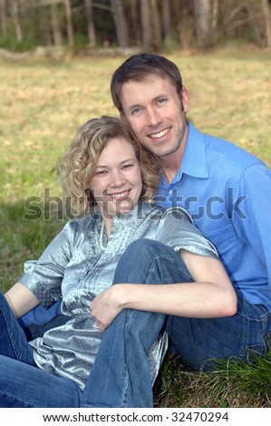 Couple smiles happily while relaxing outdoors.  Both are wearing jeans.  Man is wearing a blue shirt and woman a silk grey blouse.