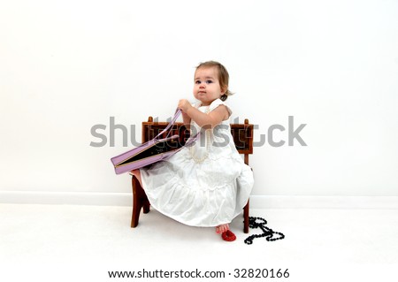 Adorable baby pulls necklaces and bracelets from mom\'s purse.  She is sitting on a stool and wearing a white long dress and hairbow.  She is barefoot.  Solid white room for background.