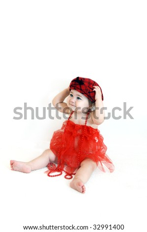 Adorable infant sits in a red tutu and doffs a red tam in red check with black bow.  She is wearing a string of red beads and a bracelet lays at her feet.