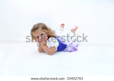 A tired little ballerina lays on the floor in an all white room.  She is dressed in a ballerina costume of lilac and purple.  Child is grinning and resting her head in her hands.
