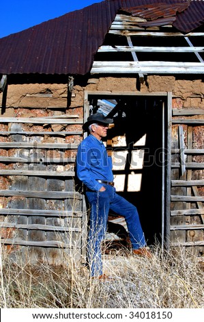 Attractive male, dressed in denim and boots, leans against the door jam of an old adobe home outside of Alberquerque, New Mexico.
