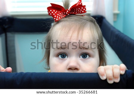 Baby girl stands at her play pen rim and begs with beseeching eyes to be lifted out of her captivity.  She has a red polka dot ribbon in her hair.
