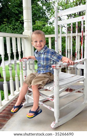 Small boy grins as he sits on a weathered rocking chair on the front porch of his home.  He is wearing flip flops, shorts and short sleeved shirt.